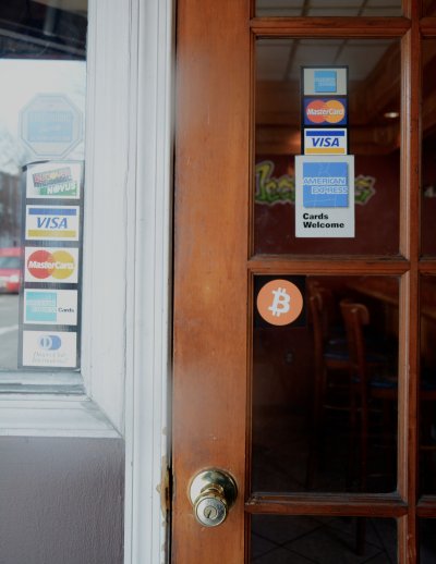 The Bitcoin logo, pictured above on the door glass, sits below traditional credit card stickers posted on the doorway at the Baltimore bar, Bad Decisions. (Photo: Patrick Farrell)