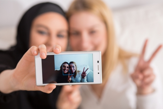 Young muslim woman doing selfie with catholic friend. (Stock photo)