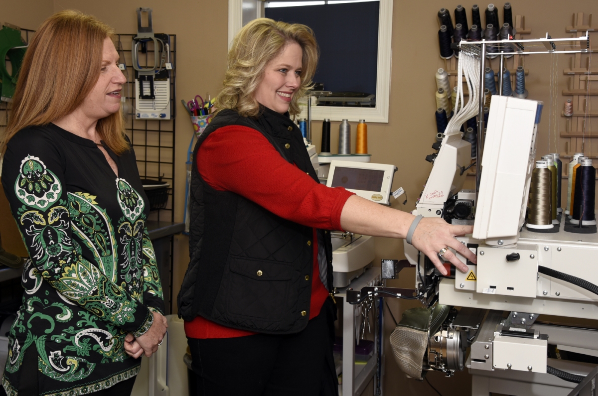 SBDC Consultant Kathy MacAdams, left, watches Ali Banholzer demonstrate the embroidery machine at Wear Your Spirit Warehouse, Banholzer’s business in Huntingtown. (Submitted photo)