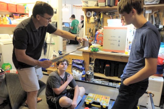 Dan Ensminger, left, head coach of the Bot Brigade Quartic robotics team, explains a practical application of building robotics to two team members in his garage, while other team members work out 3D modeling around from his kitchen table. Ensminger has led local Bot Brigade teams for the past 14 years. (Photo courtesy of Bot Brigade Quartic)